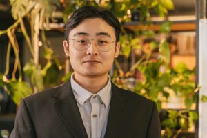Portrait of a man in a suit with eyeglasses, set against a leafy indoor background.